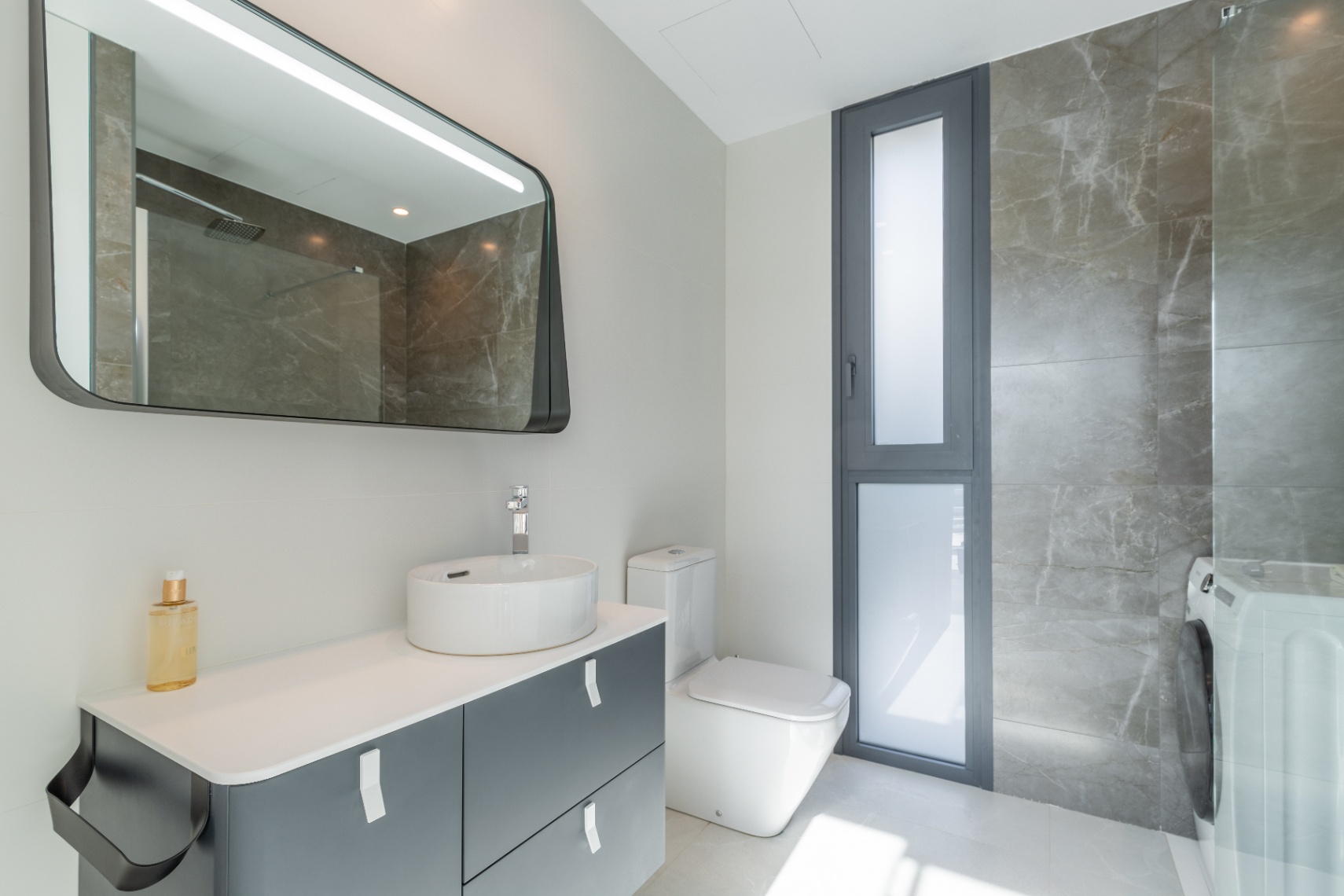 Modernes Badezimmerdetail mit Keramikwaschbecken, hinterleuchtetem Spiegel und grauer Marmorverkleidung.
