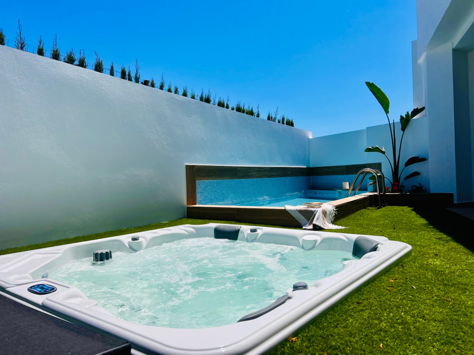 Außen-Whirlpool in Kunstrasen eingebettet, neben einem privaten Pool mit blauen Fliesen und hohen weißen Mauern für maximale Privatsphäre.