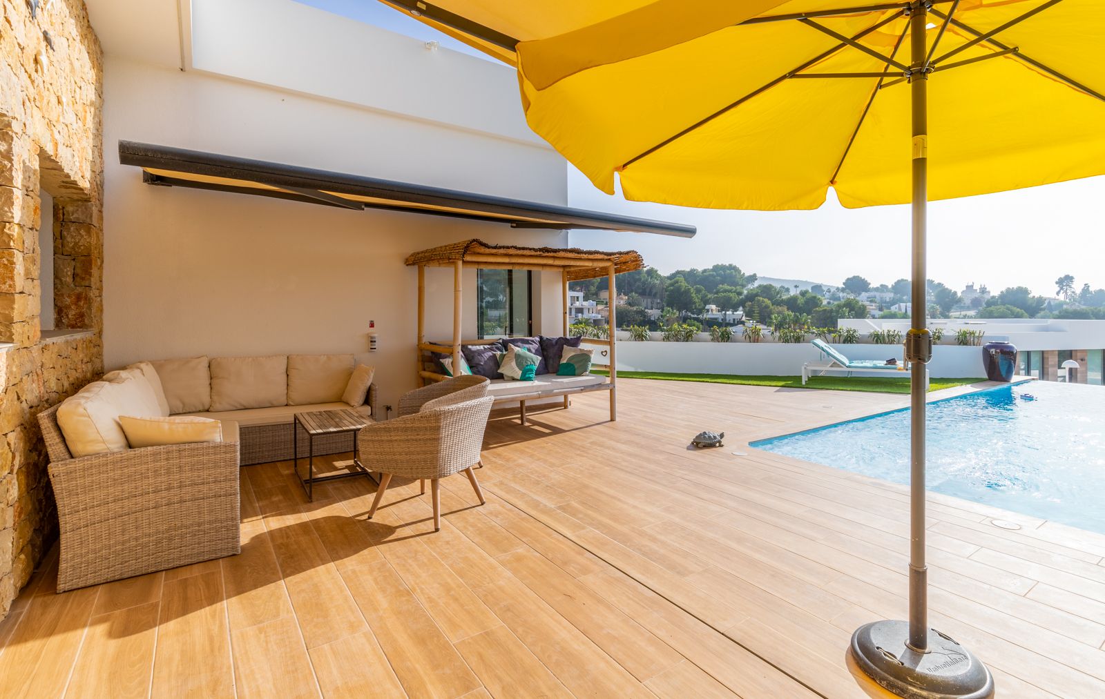 Außenterrasse mit Feinsteinzeug in Holzoptik, Korbmöbel-Lounge, Bambus-Daybed und Poolbereich, ergänzt durch eine Natursteinwand und einen gelben Sonnenschirm.