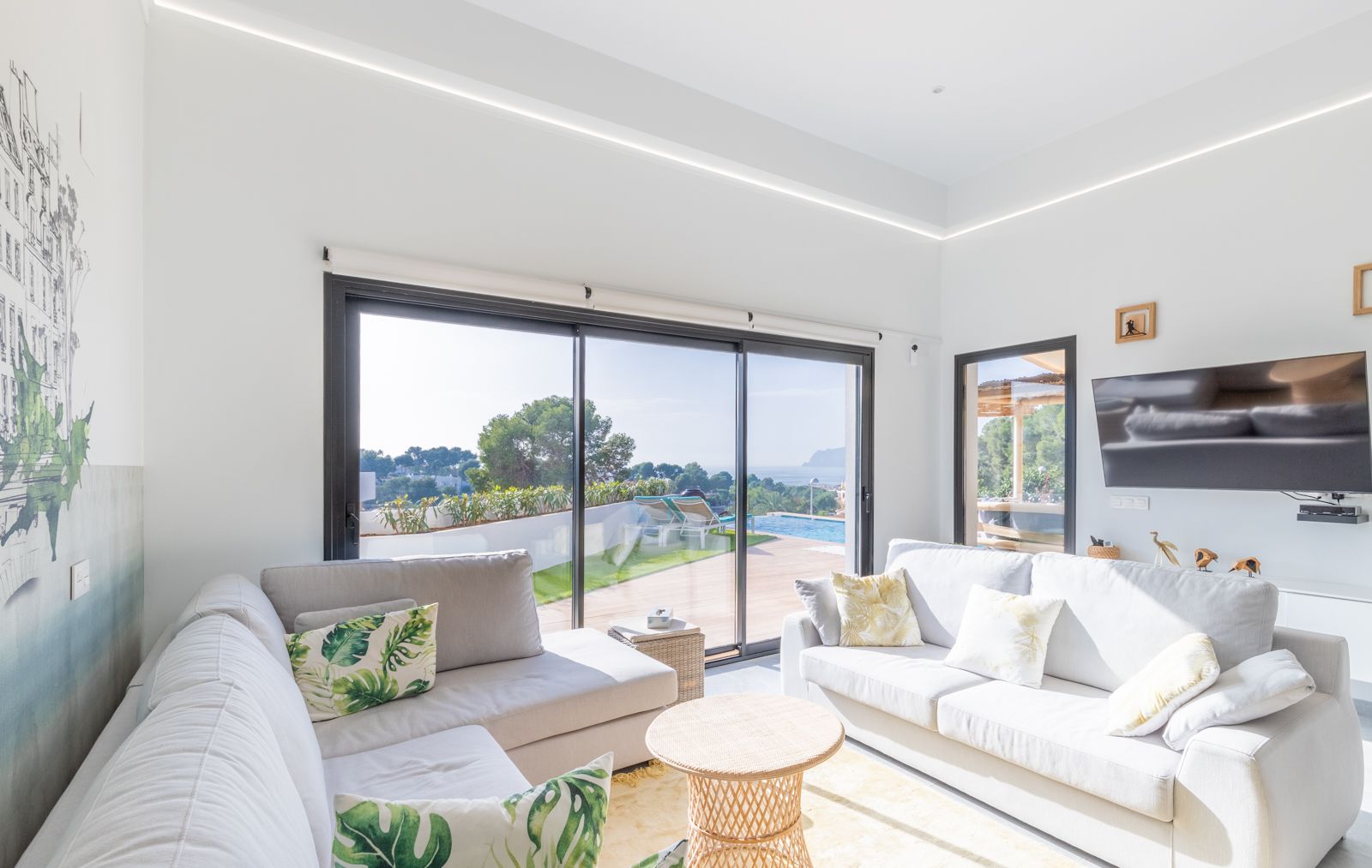 Heller Wohnbereich mit grauen Sofas, integrierter LED-Deckenbeleuchtung und großen Glasschiebetüren, die den Blick auf den Pool und das Meer freigeben.