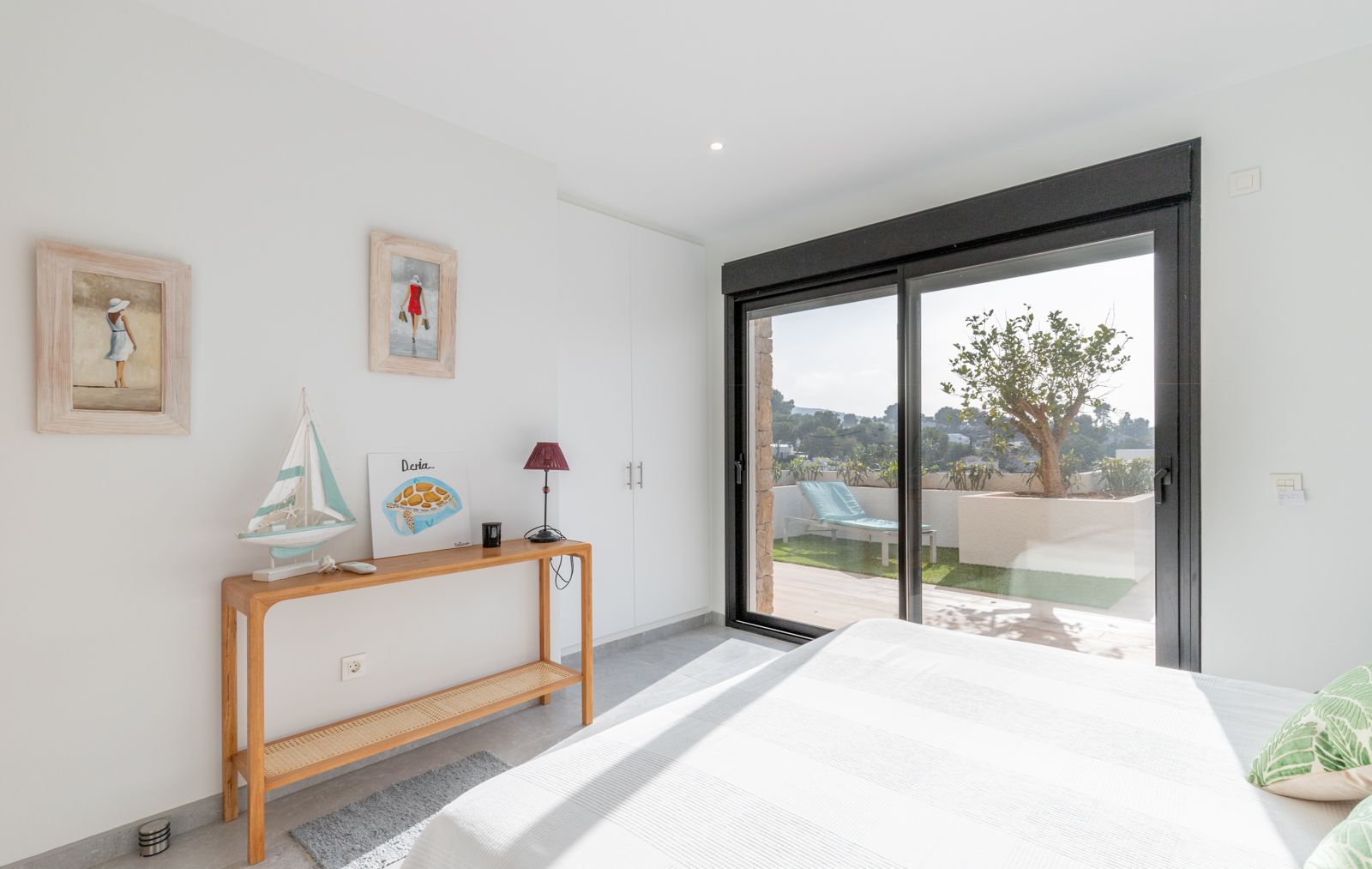 Helles Schlafzimmer mit weißen Einbauschränken und Glasschiebetüren, die auf eine private Terrasse mit Kunstrasen und Sonnenliege führen.
