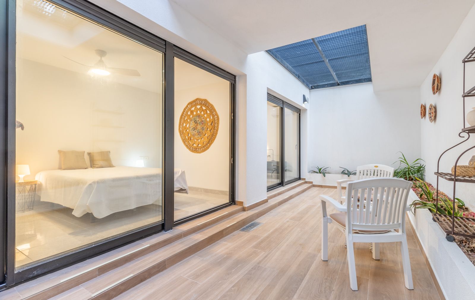 Innenhof mit Bodenfliesen in Holzoptik, Sitzbereich und großen Glasschiebetüren, die in ein helles, minimalistisch gestaltetes Schlafzimmer führen.