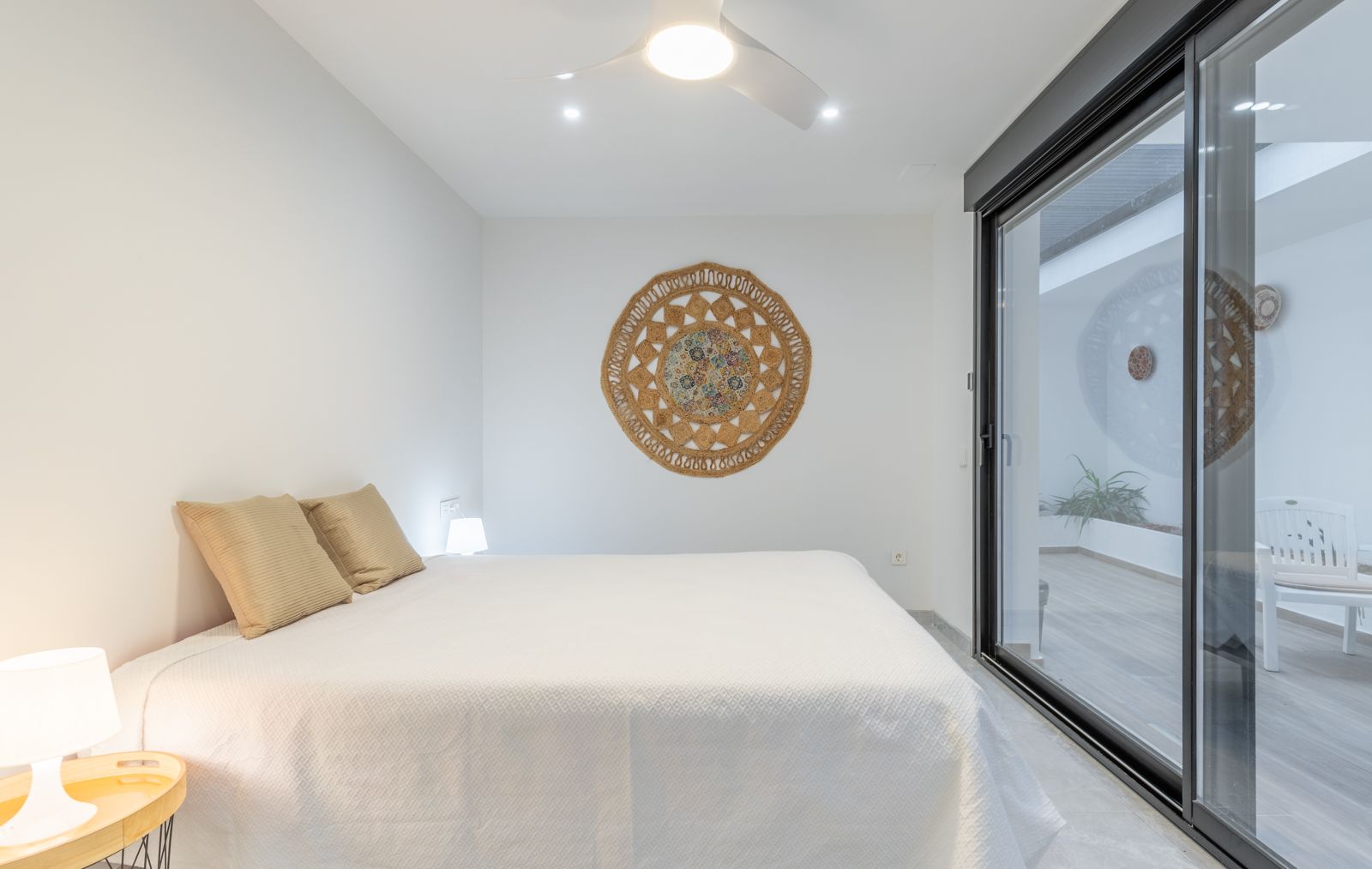 Minimalistisches Schlafzimmer mit direktem Zugang zur Terrasse durch große Glasschiebetüren, Deckenventilator und Wanddekoration aus Naturfasern.