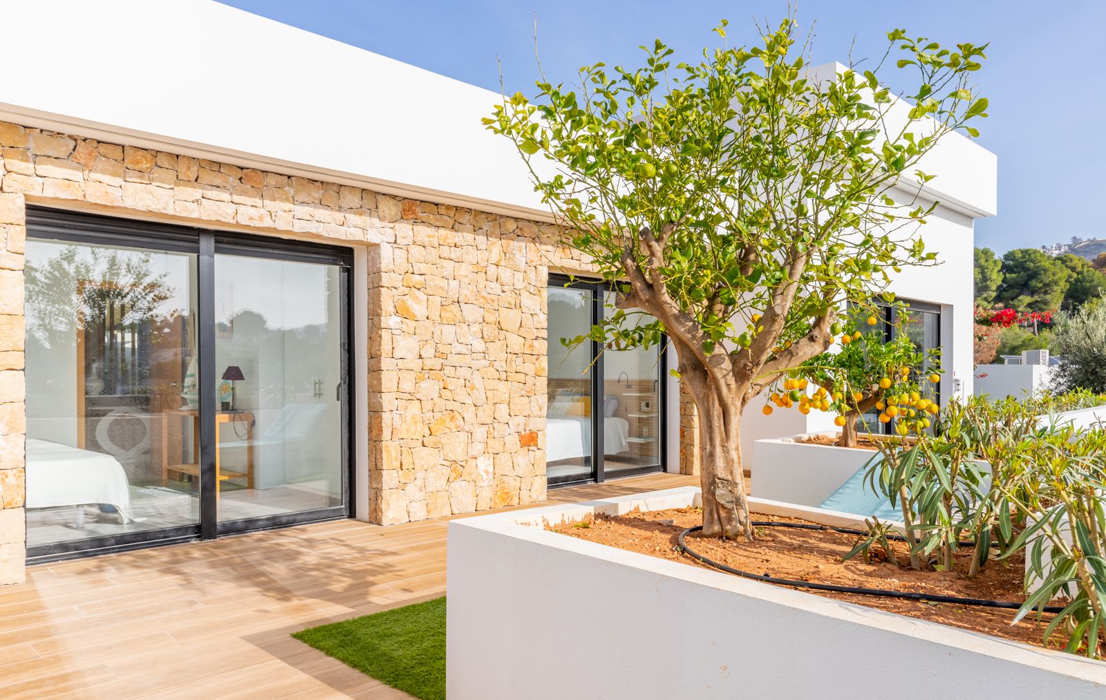 Moderne Fassade mit Natursteinwänden und bodentiefen Glasschiebetüren, die auf eine Terrasse mit integrierten Pflanzkübeln und einem Zitronenbaum führen.