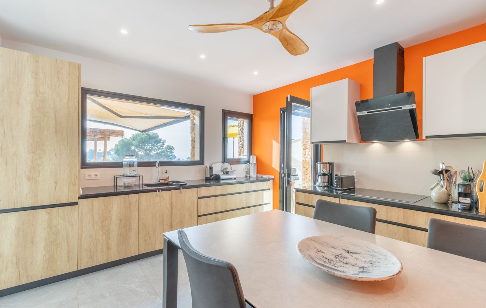 Moderne Küche mit hellen Holzfronten, schwarzen Arbeitsplatten und einer orangefarbenen Wand. Ausgestattet mit einem Holz-Deckenventilator und Blick auf die Terrasse.