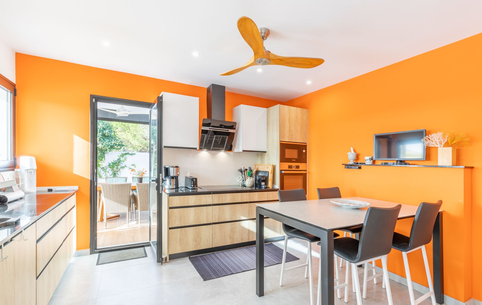 Moderne Küche mit leuchtend orangefarbenen Wänden, hellen Holzschränken und integriertem Essbereich mit direktem Zugang zur Außenterrasse.