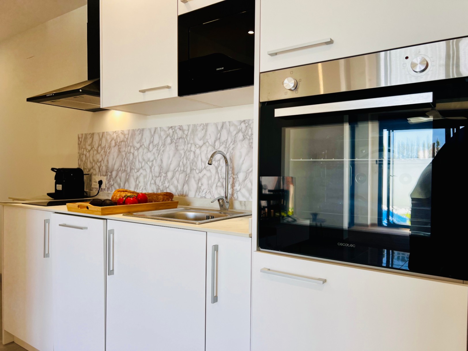 Moderne Küche mit weißen Schränken, integrierten Cecotec-Geräten und einer Rückwand in Marmoroptik. Ausgestattet mit Edelstahlspüle und schwarzer Dunstabzugshaube.