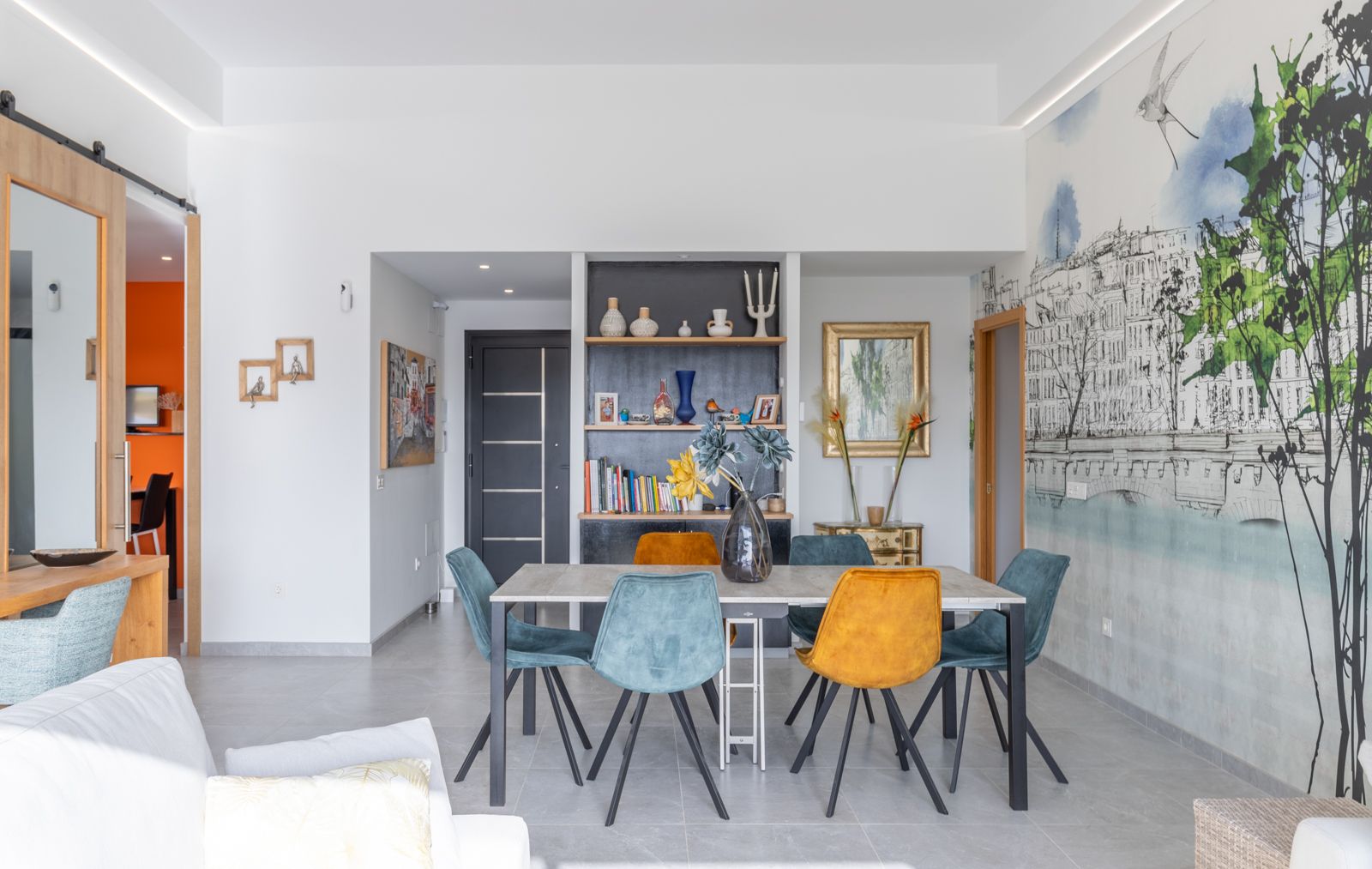 Moderner Essbereich mit minimalistischem Tisch und Samtstühlen in Blau und Ocker. Ein künstlerisches Wandbild und integrierte Regale mit LED-Beleuchtung prägen den Raum.