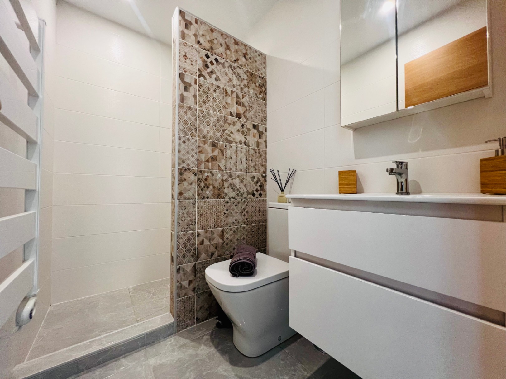 Modernes Badezimmer mit ebenerdiger Dusche, dekorativer Akzentwand mit geometrischen Fliesen, weißem Waschtisch und Handtuchheizkörper.