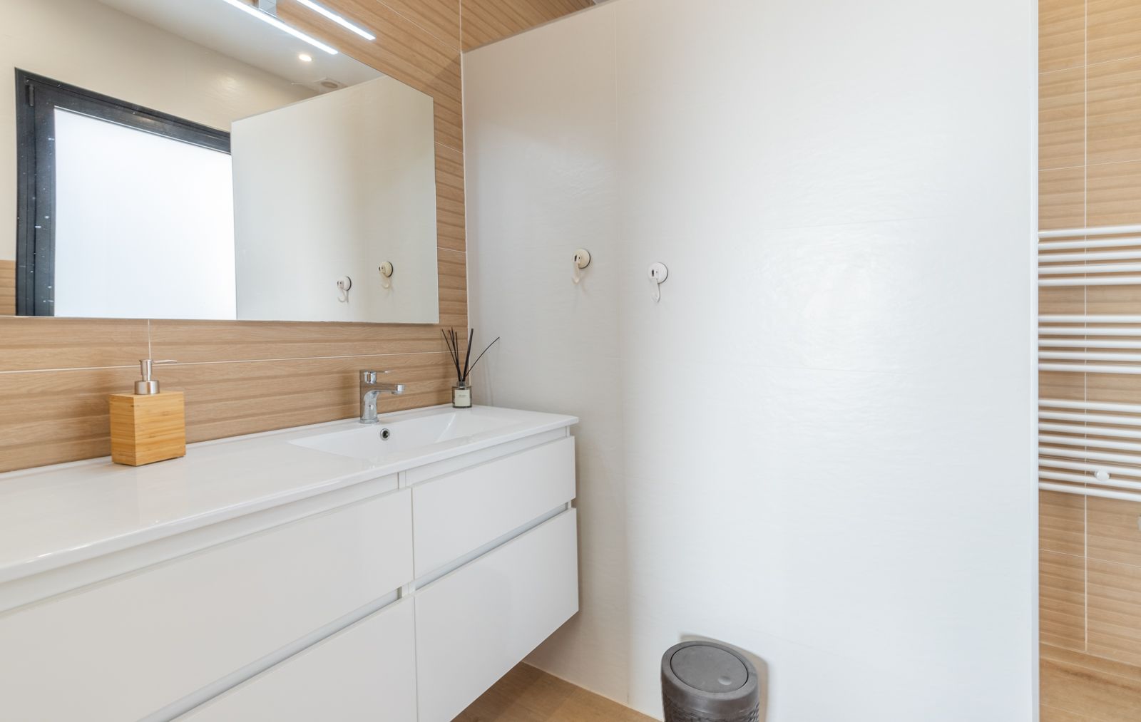 Modernes Badezimmer mit schwebendem weißem Waschtisch, Wandfliesen in Holzoptik, großem Spiegel mit LED-Beleuchtung und einem weißen Handtuchheizkörper.