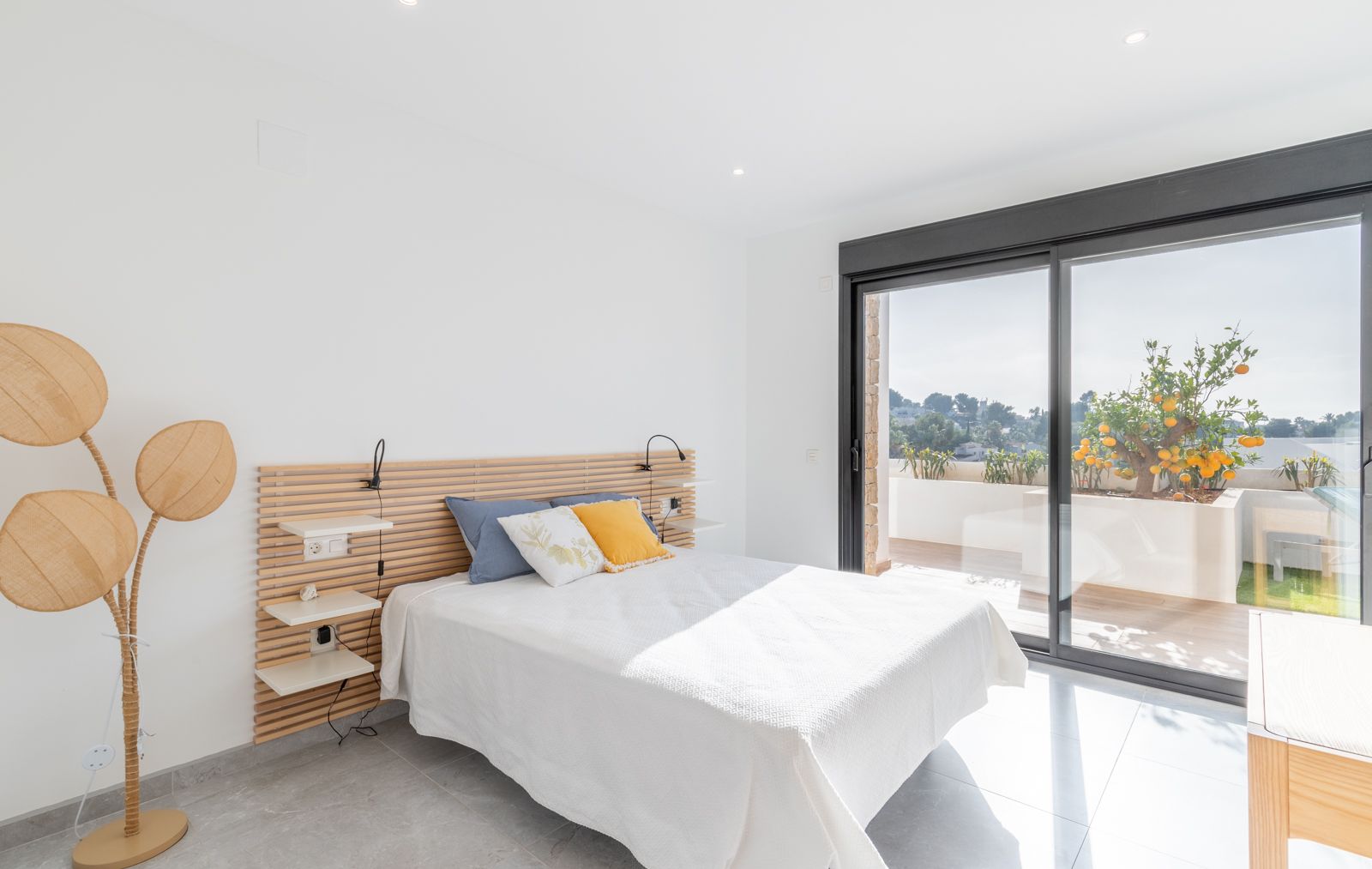 Modernes Schlafzimmer mit Holzlatten-Kopfteil, integrierten Regalen und großen Glasschiebetüren, die auf eine Terrasse mit Zitrusbäumen führen.