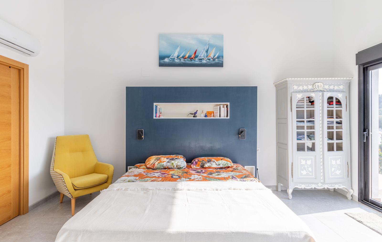 Modernes Schlafzimmer mit integriertem blauem Kopfteil, gelbem Sessel und klassischem Schrank. Inklusive Klimaanlage und direktem Zugang nach draußen durch Glasschiebetüren.