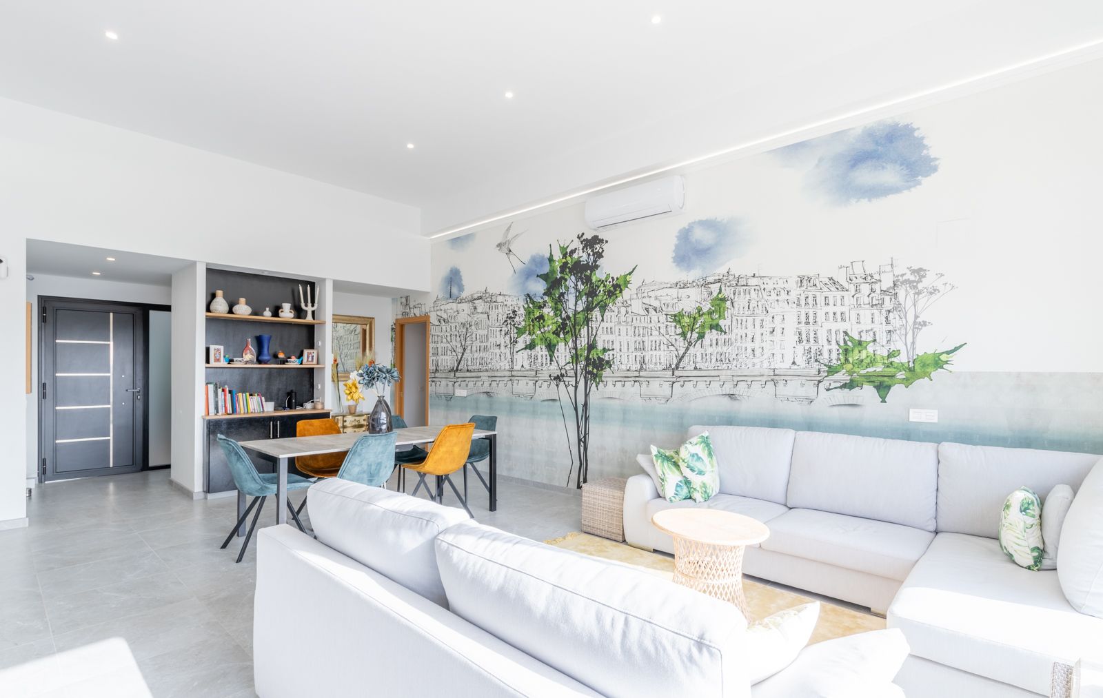 Offener Wohn- und Essbereich mit großem Ecksofa, einer dekorativen Wandmalerei im Stadtdesign und modernen Möbeln auf hellen Fliesenböden.