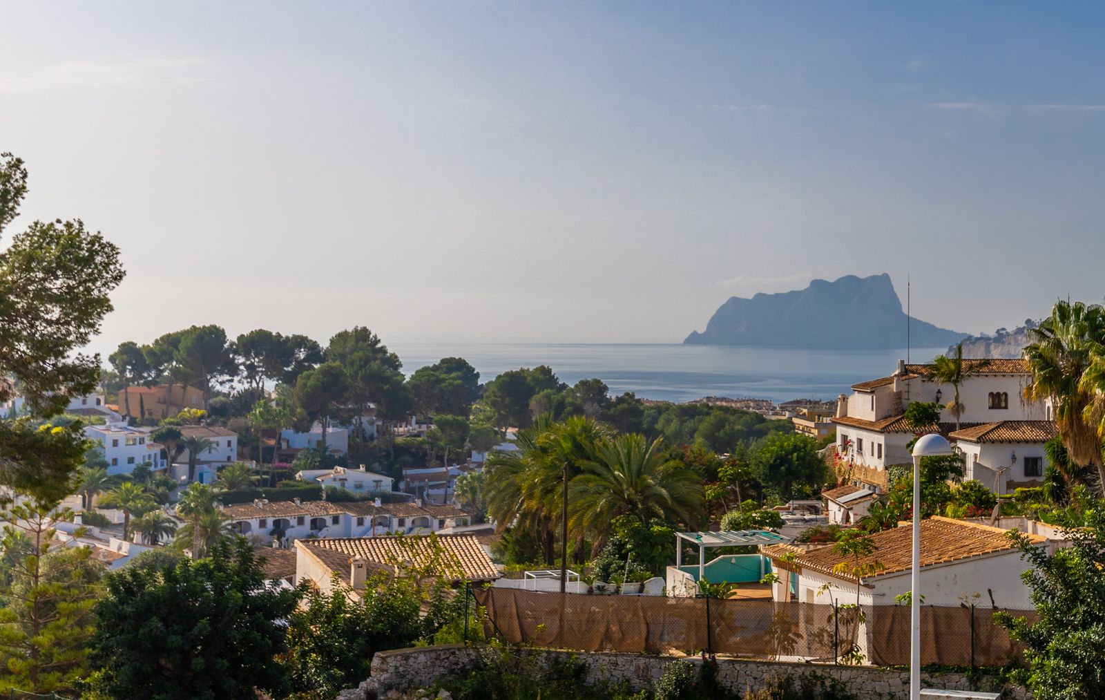 Panoramablick auf das Mittelmeer und den Peñón de Ifach, über eine Landschaft aus traditionellen weißen Villen und üppigen Palmen.