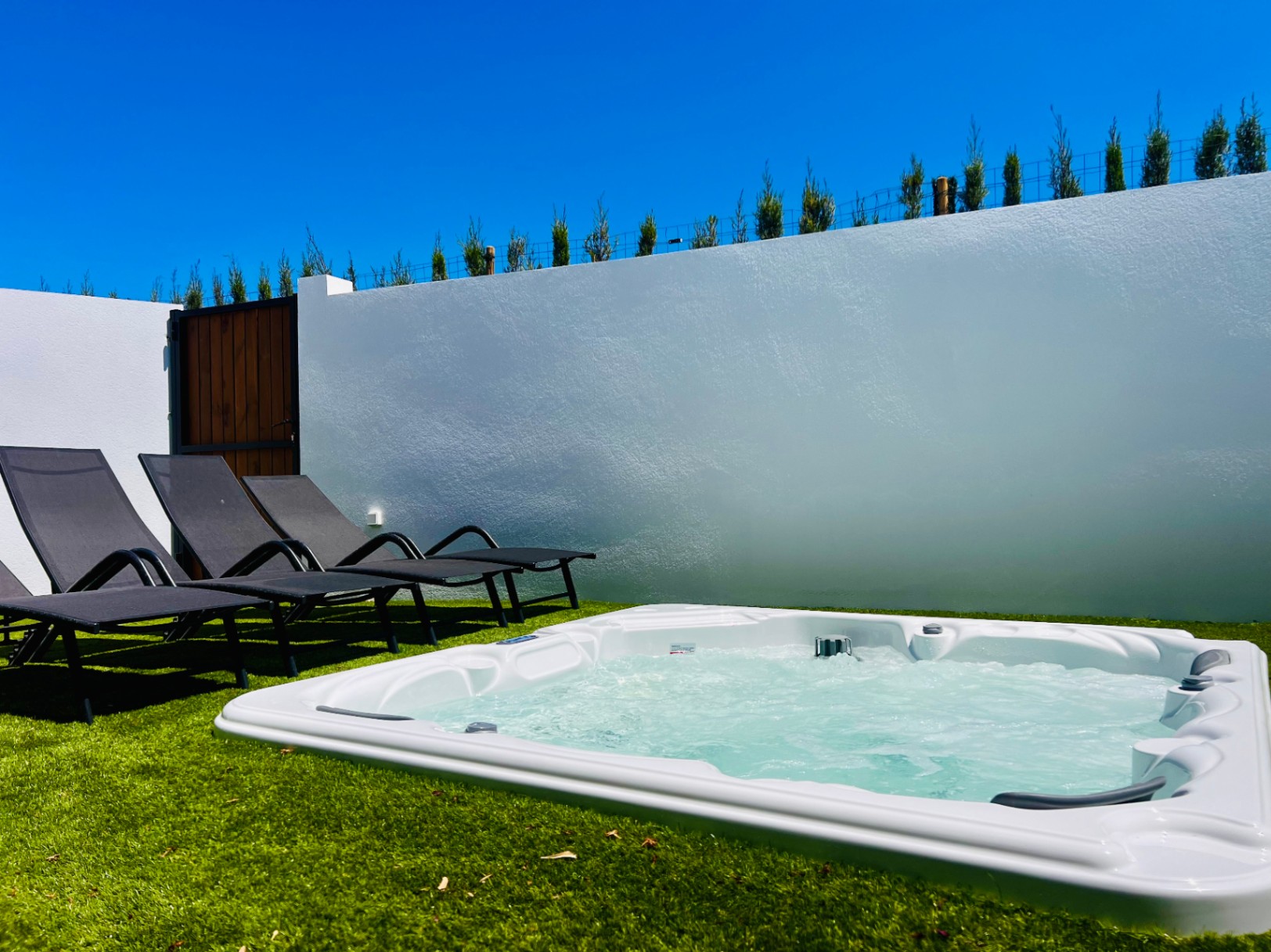 Privater Whirlpool, eingelassen in Kunstrasen, mit modernen Sonnenliegen und einer weißen Sichtschutzmauer für maximale Privatsphäre im Außenbereich.