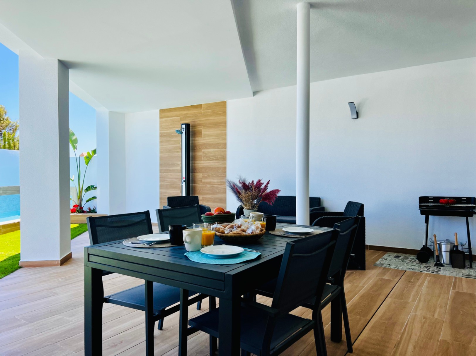 Überdachte Terrasse mit Feinsteinzeug in Holzoptik, Essbereich im Freien, Grillstation und Außendusche mit dekorativem Wandpaneel.