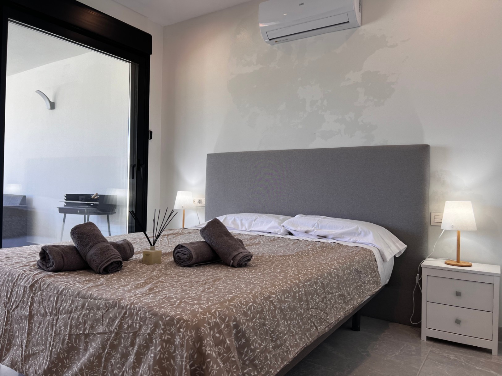 Zeitgenössisches Schlafzimmer mit grauem Stoffkopfteil, Split-Klimaanlage und direktem Terrassenzugang durch große schwarz gerahmte Schiebetüren.