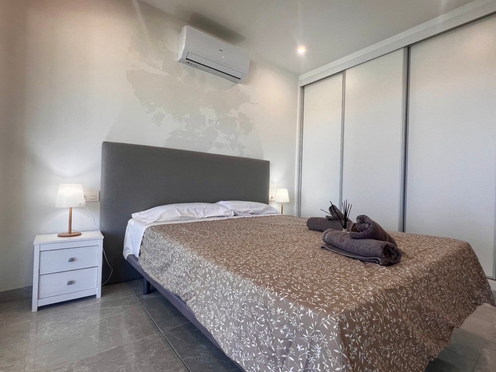 Zeitgenössisches Schlafzimmer mit weißem Einbauschrank, Klimaanlage und gepolstertem Kopfteil auf hochwertigem grauen Fliesenboden.