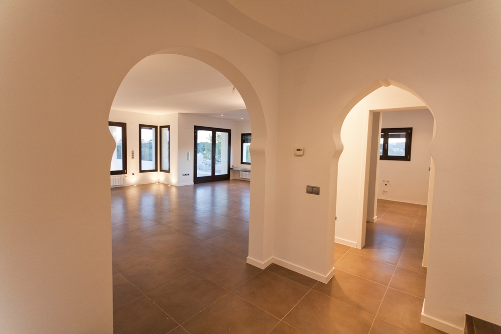 Flur mit mediterranen Rundbögen, Bodenfliesen in Terrakotta-Optik und Blick in das helle Wohnzimmer mit dunklen Aluminium-Fensterrahmen.