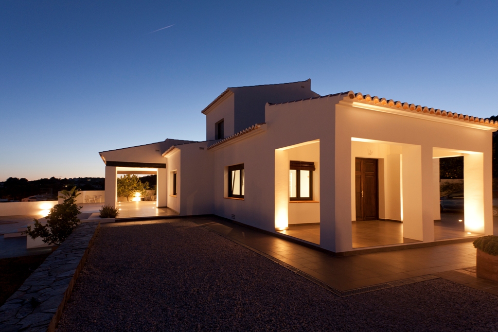 Mediterrane Fassade in der Abenddämmerung mit Bodenstrahlern an den Säulen, die die restaurierte Holzeingangstür und die weißen Außenwände betonen.