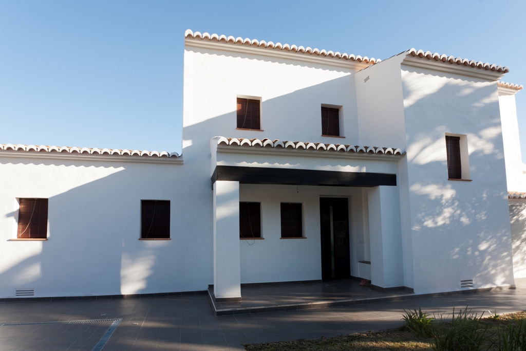 Mediterrane Fassade mit weißen Wänden, Fensterrahmen in Holzoptik und traditionellem Ziegeldach über einem schattigen Eingangsbereich mit dunklen Holzbalken.