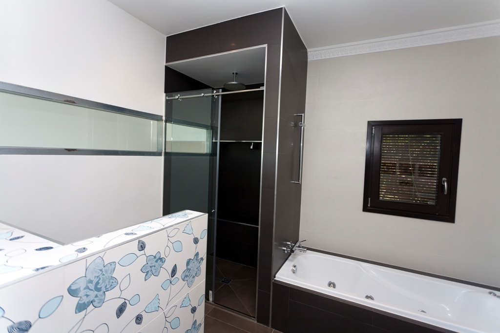 Modernes Badezimmer mit Einbaubadewanne und separater Dusche. Dunkle Akzentfliesen treffen auf helle Wände und eine dekorative Trennwand mit blauem Blumenmuster.