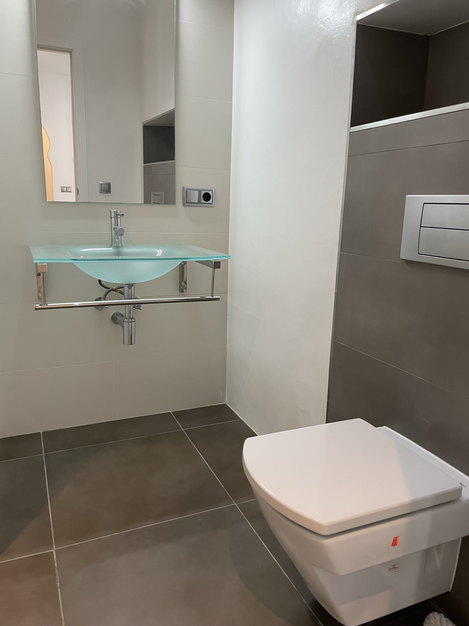 Modernes Badezimmer mit einem Waschbecken aus lichtdurchlässigem Glas und wandhängendem WC. Großformatige Fliesen in Grau und Creme sorgen für ein zeitgemäßes Design.