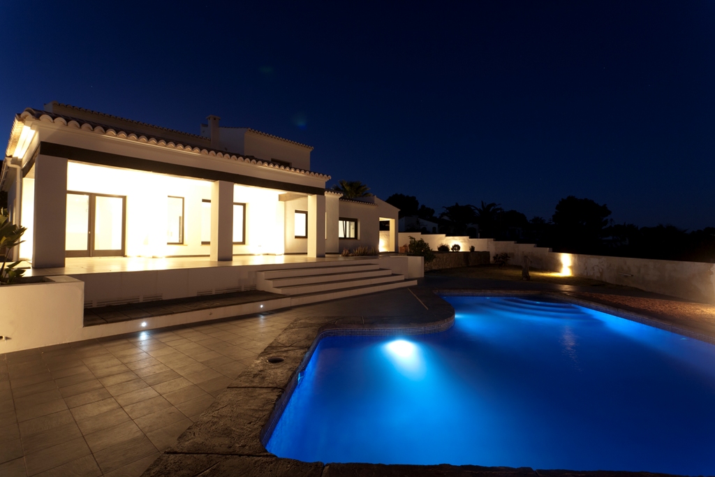 Nachtansicht der beleuchteten weißen Fassade und des unregelmäßig geformten Pools mit blauer Unterwasserbeleuchtung auf einer Fliesenterrasse.