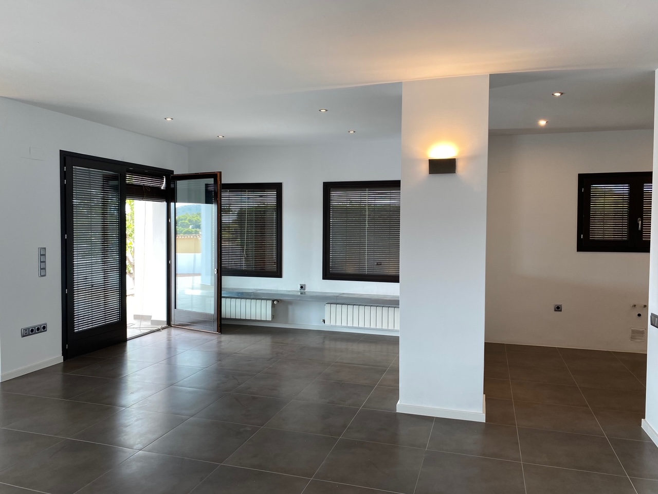 Offener Wohnbereich mit dunklen Bodenfliesen, Deckenspots und Wandheizkörpern. Die Fenster sind mit integrierten Jalousien ausgestattet und lassen viel Tageslicht herein.