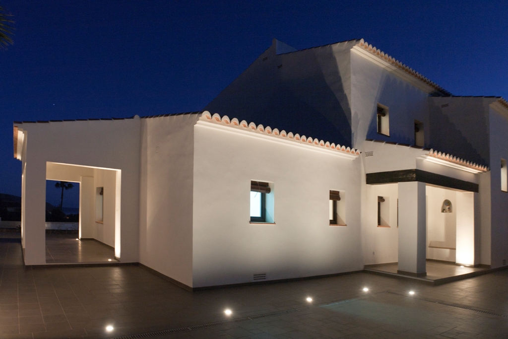 Weiße mediterrane Fassade, die bei Dämmerung durch Bodeneinbaustrahler beleuchtet wird. Mit traditionellen Dachziegeln und einer überdachten Veranda mit dunklen Holzbalken.