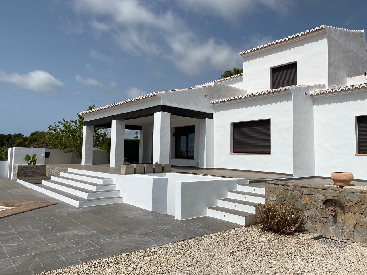 Weiße mediterrane Fassade mit dunklen Holzbalken, großzügiger Fliesenterrasse und Natursteinmauern. Inklusive Außendusche und integrierten Zugangsstufen.