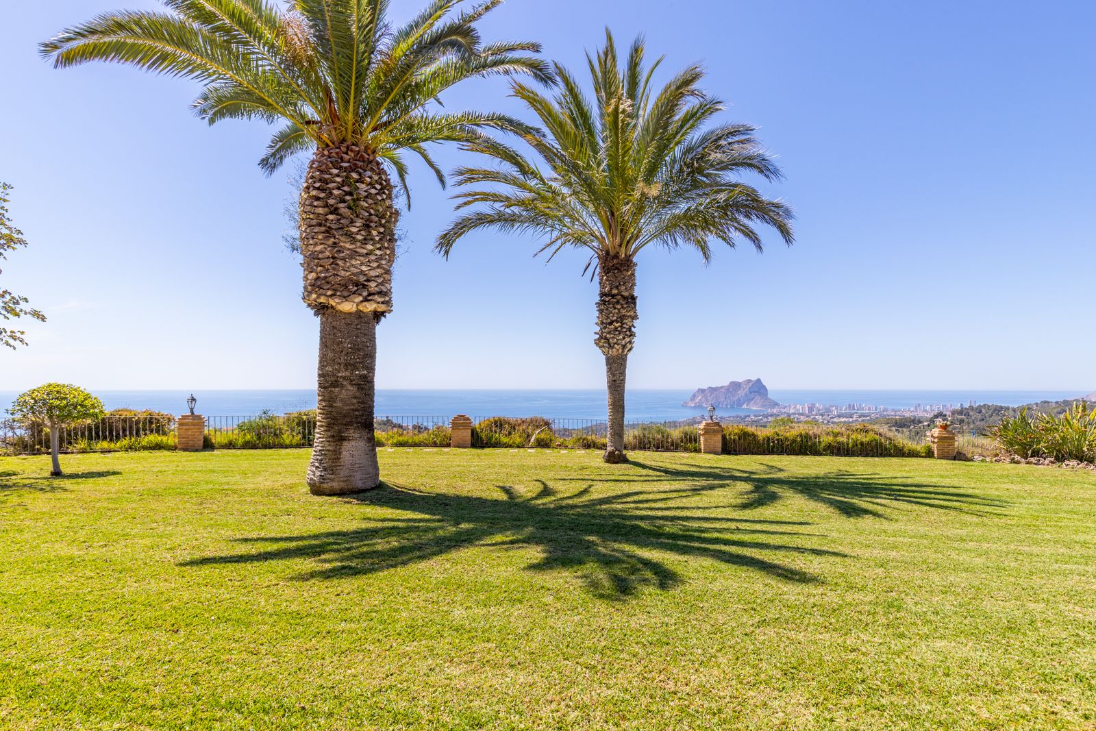 Blick auf den Garten mit grünem Rasen, Palmen und Blick auf das Mittelmeer und den Peñón de Ifach.