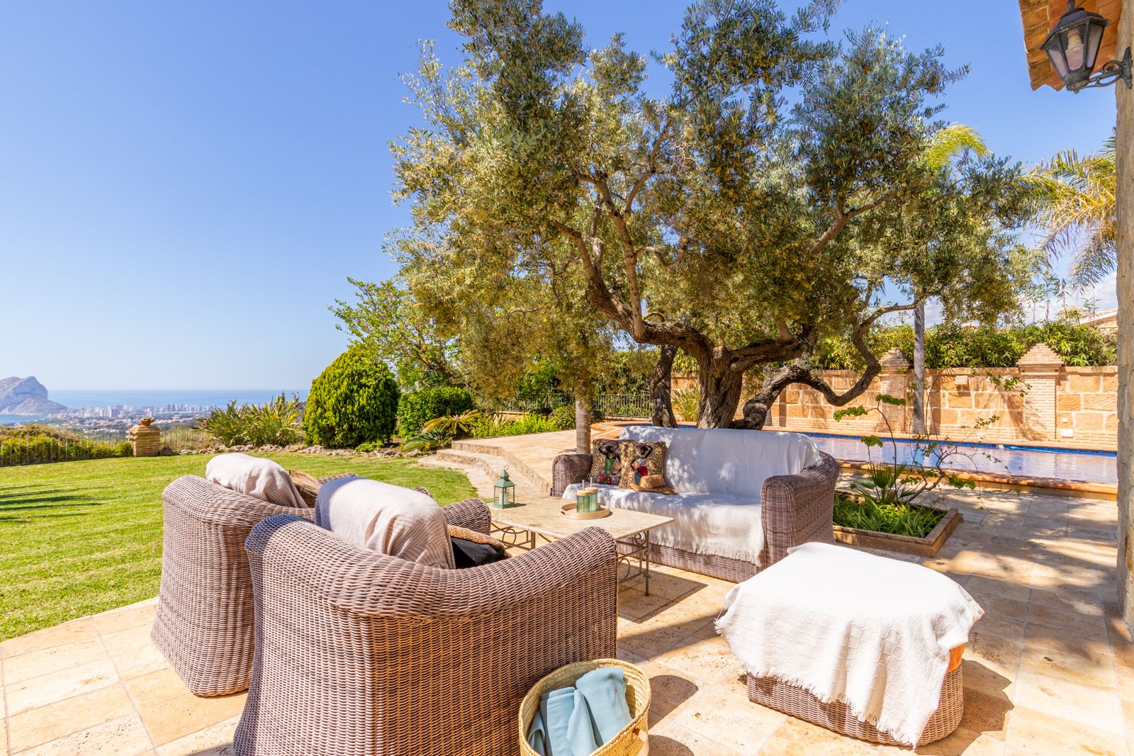 Terrasse mit Korbmöbeln und Meerblick, beschattet von einem alten Olivenbaum.
