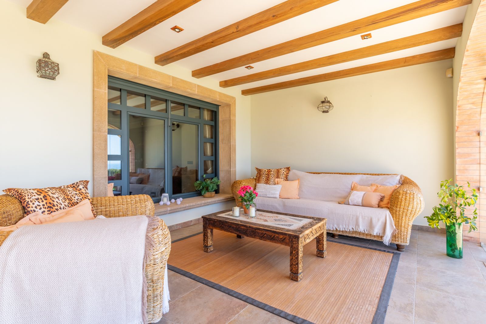 Überdachte Veranda mit Holzbalken, Korbcouch und Sessel sowie geschnitztem Holztisch.