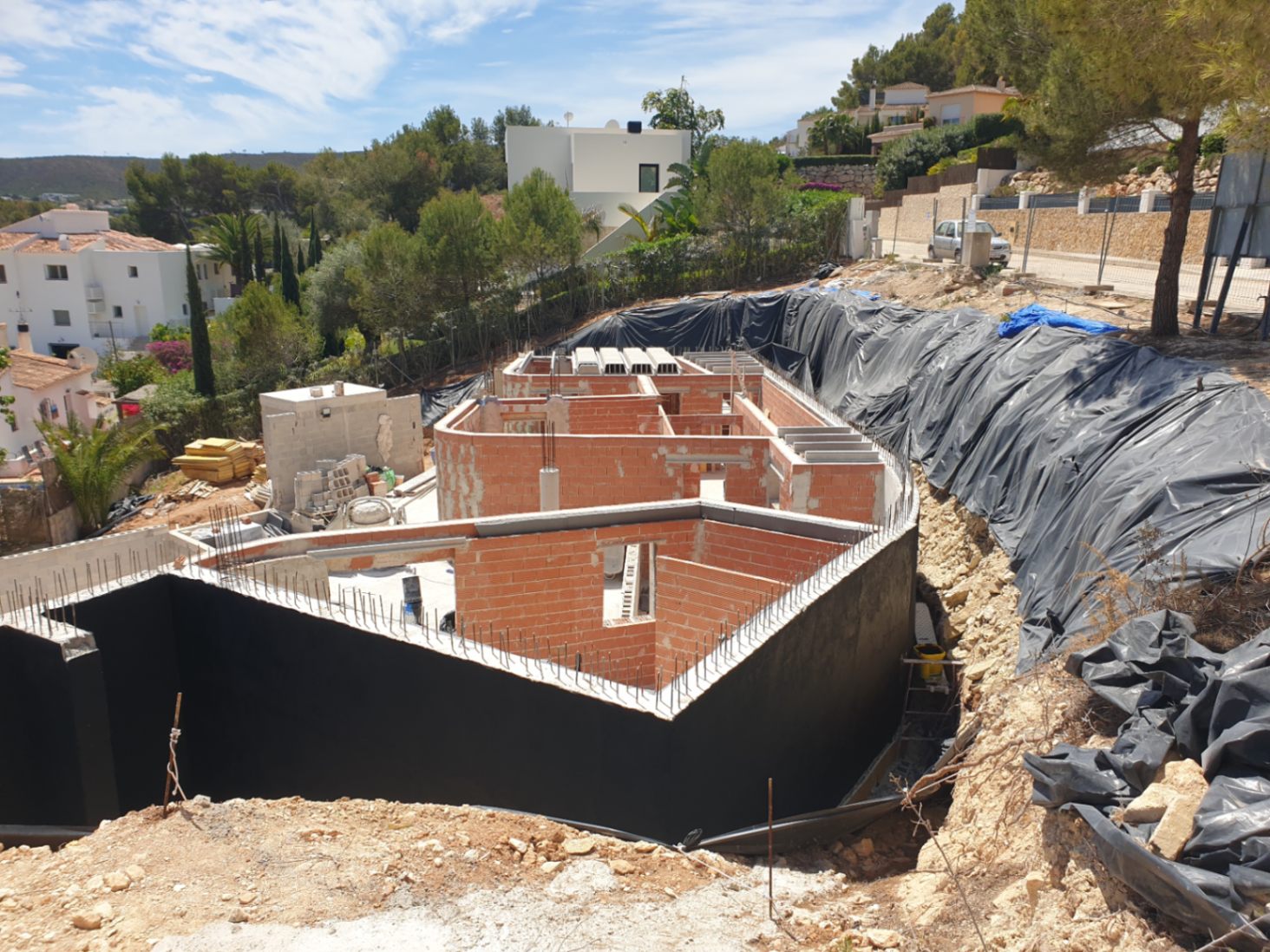 Aktueller Baustatus der Villa mit Ziegelmauerwerk und abgedichteten Stützwänden auf einem Hanggrundstück in einer Wohngegend.