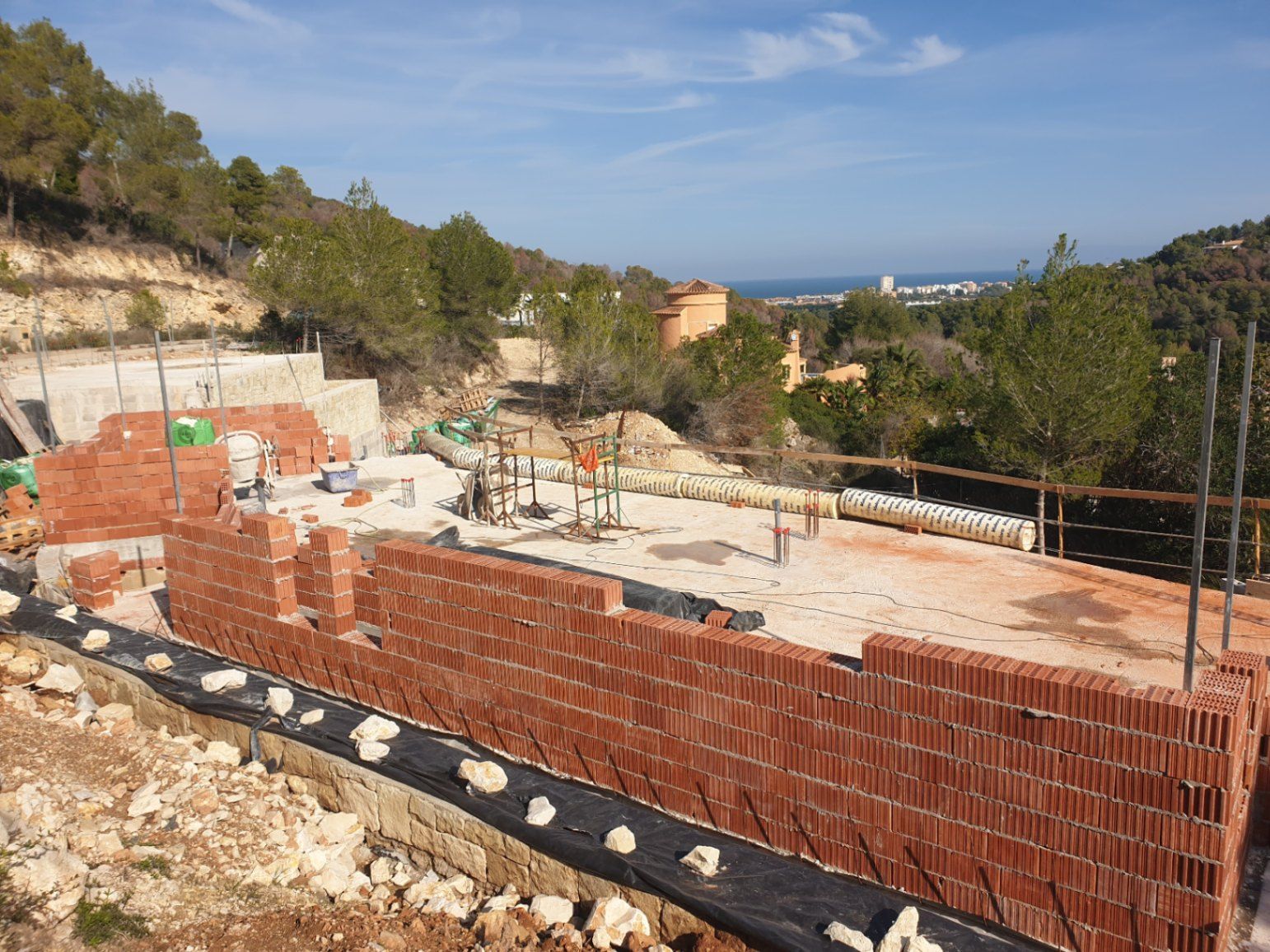 Aktueller Baustatus mit hochziehenden Ziegelwänden auf der Fundamentplatte, eingebettet in die Hügellandschaft von Jávea mit weitem Blick auf das Meer.
