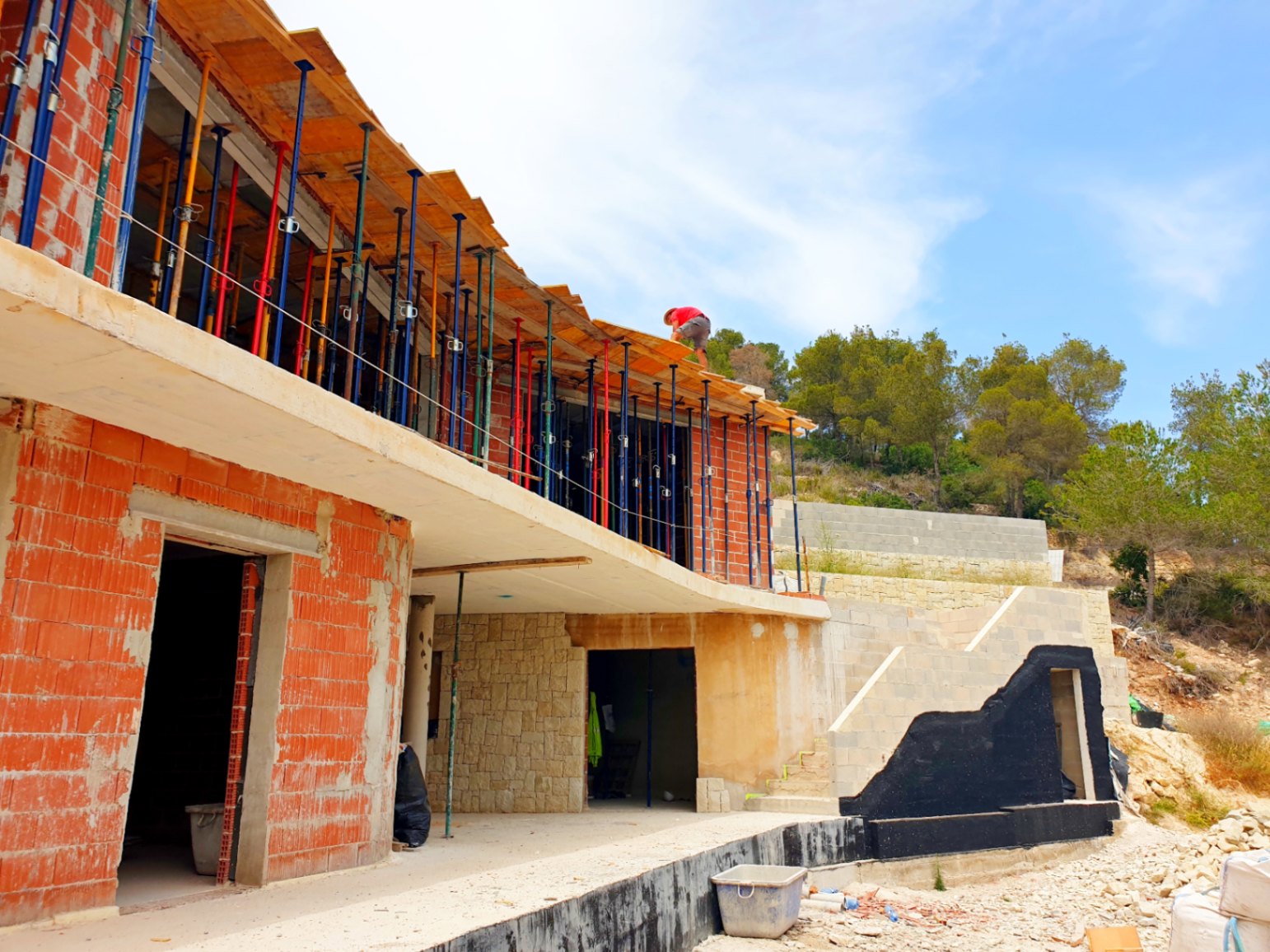 Außenansicht der Villa im Bau, mit der Ziegelstruktur, Betonplatten mit Baustützen und ersten Natursteinmauern auf dem terrassierten Grundstück.