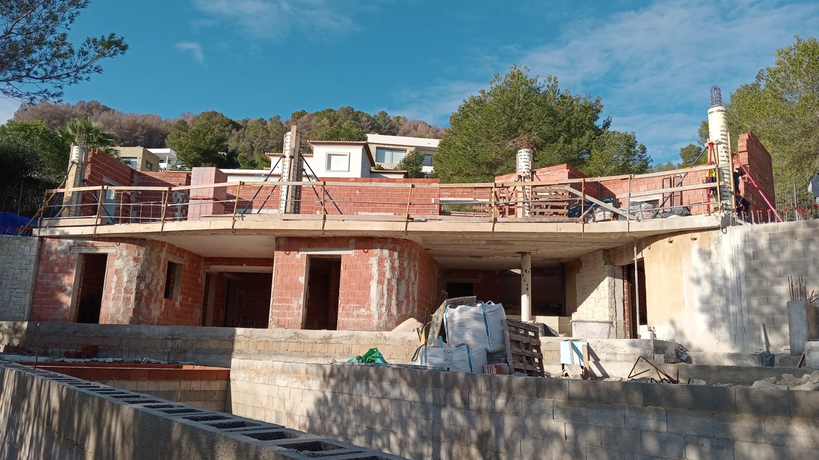 Außenansicht des Rohbaus mit der markanten geschwungenen Terrasse und den gemauerten Wohnebenen vor einer Kulisse aus Pinienbäumen.