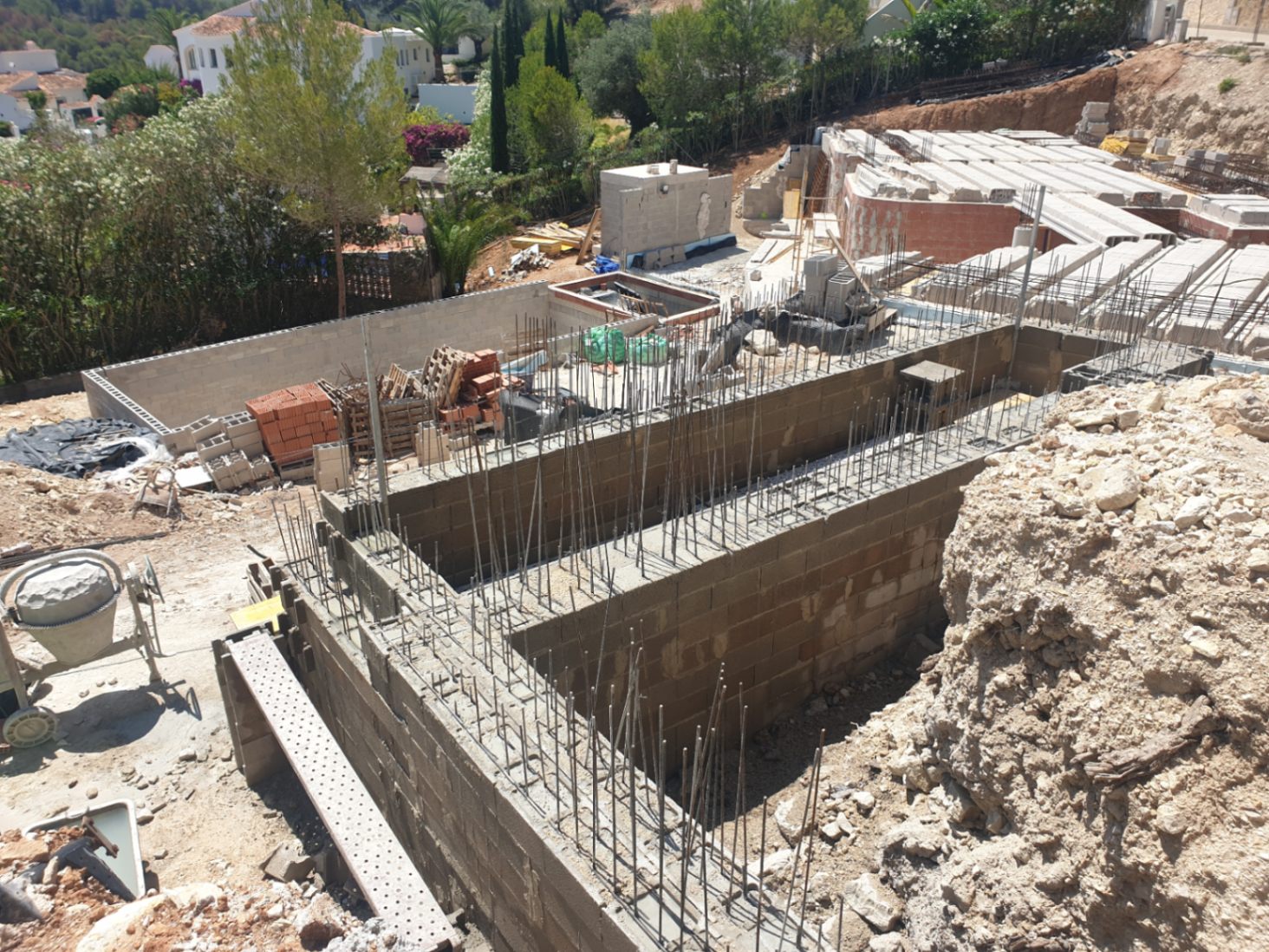 Frühe Bauphase mit Fundamentmauern, Stahlbewehrung und Betonsteinen auf einem erhöhten Grundstück inmitten mediterraner Vegetation.