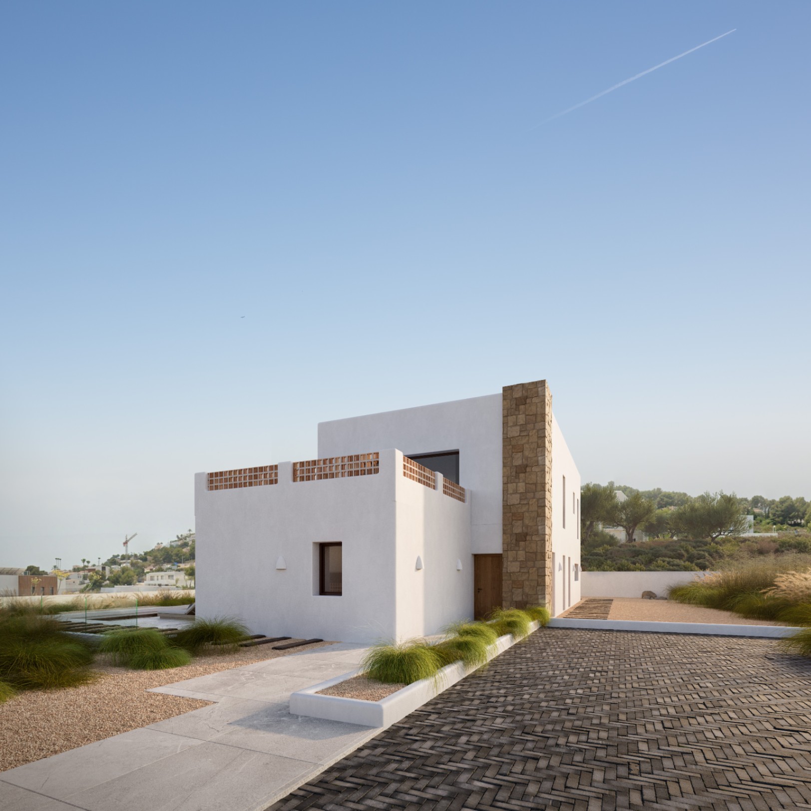 Moderne mediterrane Fassade mit weißen Wänden, einer Natursteinmauer und Terrasse mit Terrakotta-Gitter, eingebettet in eine minimalistische Gartenanlage mit Pflasterwegen.