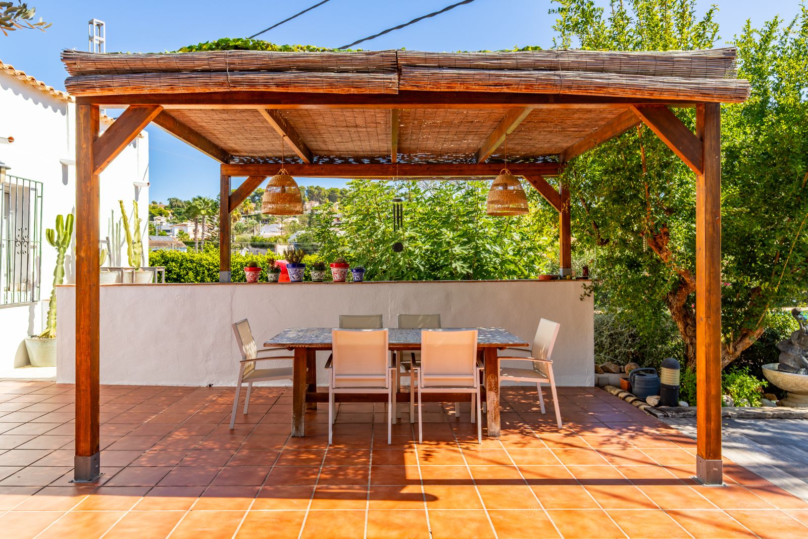 Außen-Essbereich unter einer Holzpergola mit Schilfrohr-Eindeckung, Korbpendelleuchten und einem Fliesentisch auf Terrakottaboden.