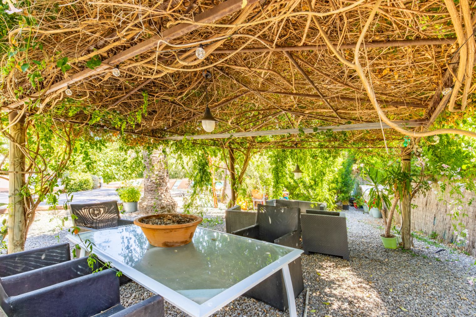 Außen-Essbereich unter einer natürlichen, dicht bewachsenen Pergola mit Glastisch, dunklen Rattansesseln und Kiesboden inmitten einer gepflegten Gartenanlage.
