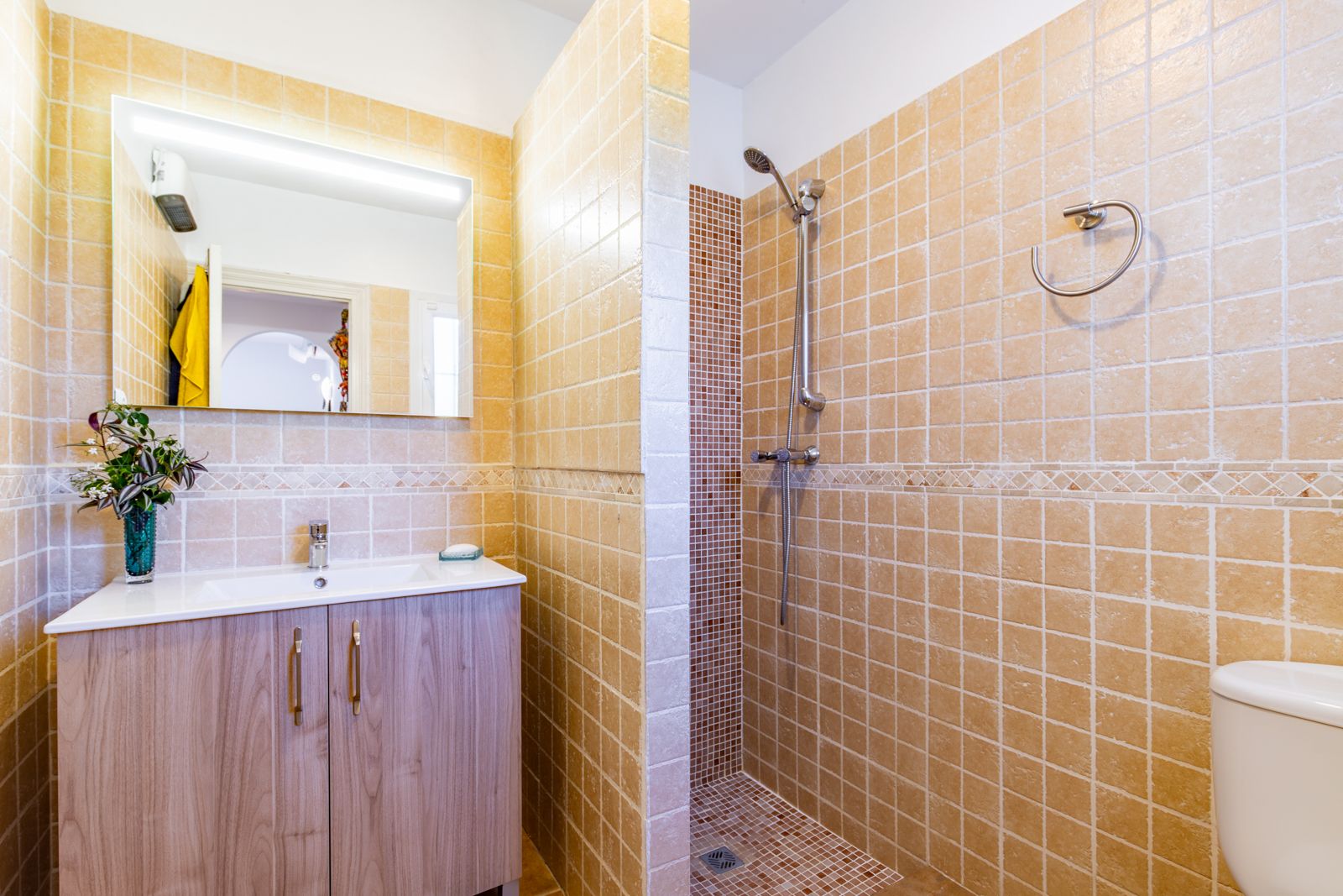 Badezimmer mit begehbarer Dusche, Mosaikboden und beigefarbenen Fliesen. Ein Waschtisch in Holzoptik und ein beleuchteter Spiegel ergänzen das Design.