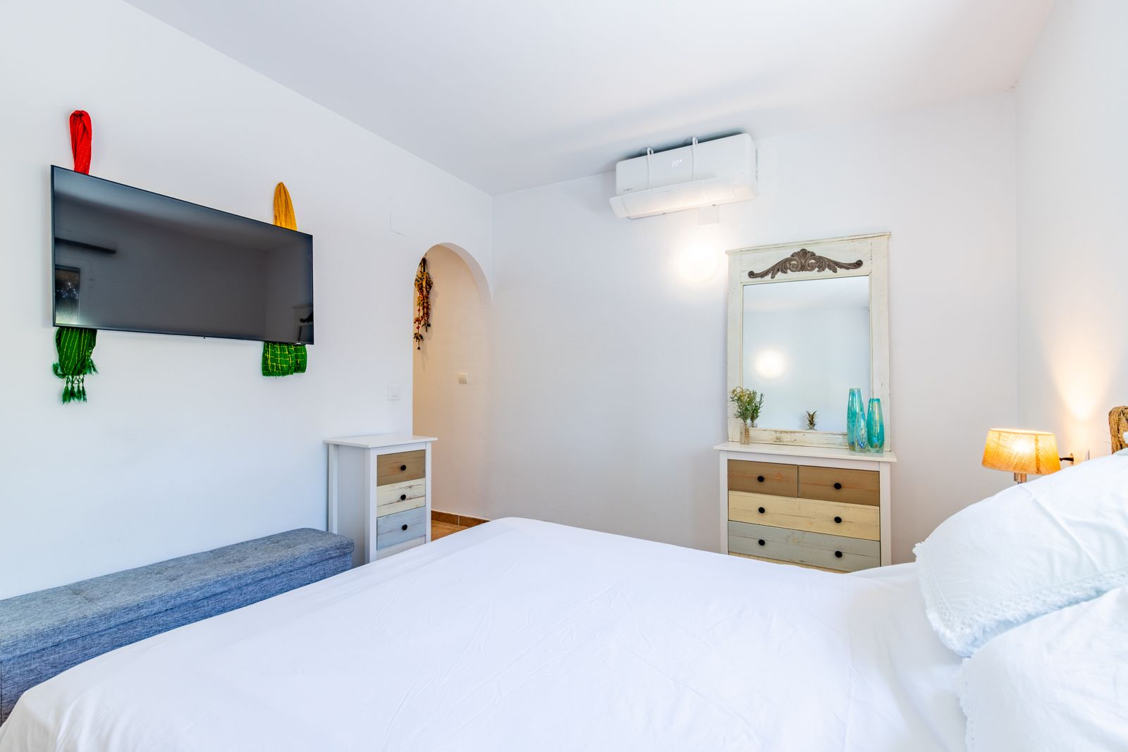 Helles Schlafzimmer mit Doppelbett, rustikalen Kommoden, Klimaanlage und wandmontiertem Fernseher in schlichtem, weißem Design.