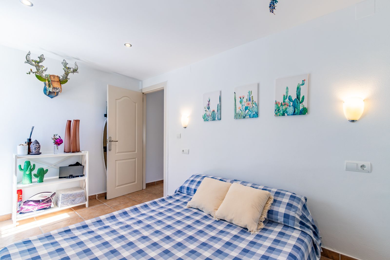 Helles Schlafzimmer mit Terrakottaboden, Bett mit blau karierter Bettwäsche und Kaktus-Dekoration. Ausgestattet mit Einbaustrahlern und warmen Wandleuchten.