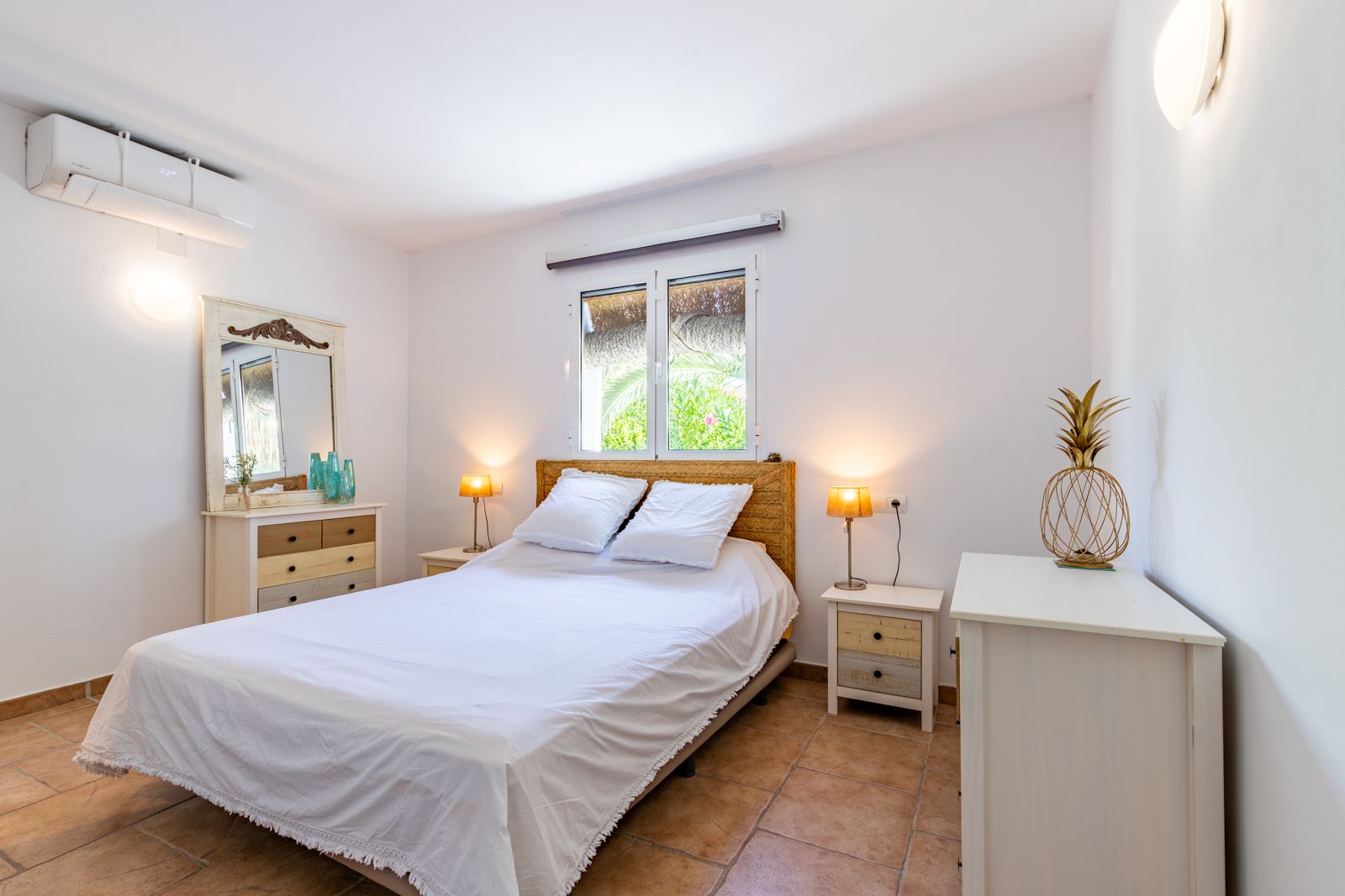 Helles Schlafzimmer mit Terrakottaboden, Klimaanlage und rustikalen Holzmöbeln. Das Zimmer bietet ein Kopfteil aus Naturfasern und ein Fenster mit Blick in den Garten.