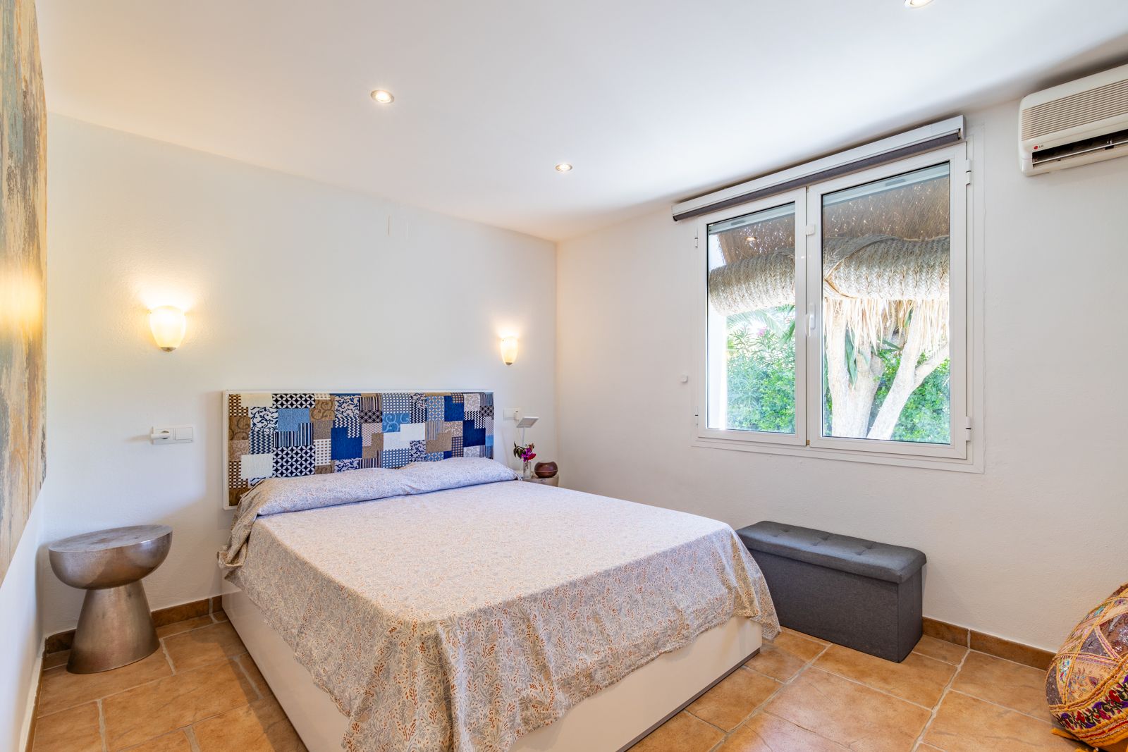 Helles Schlafzimmer mit Terrakottaboden, Patchwork-Kopfteil, Klimaanlage und Fenster mit Blick auf die Gartenanlage.