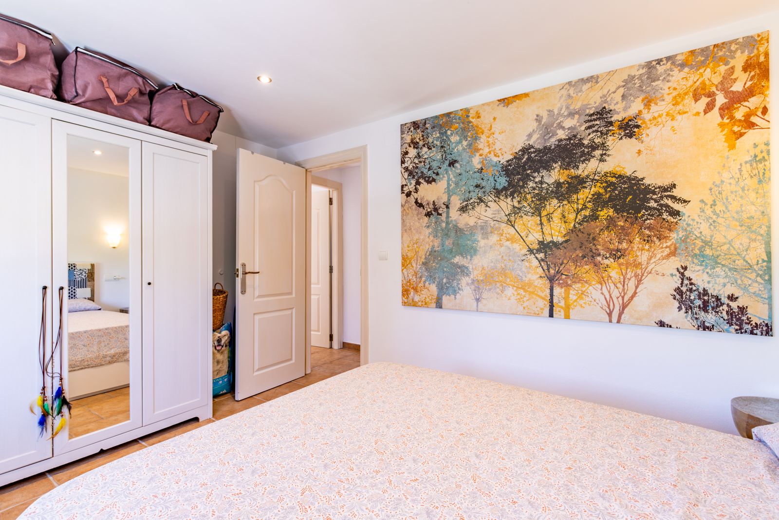 Helles Schlafzimmer mit weißem dreitürigem Kleiderschrank, Mittelspiegel und einem großen dekorativen Wandbild. Fliesenboden und dezente Deckenbeleuchtung.