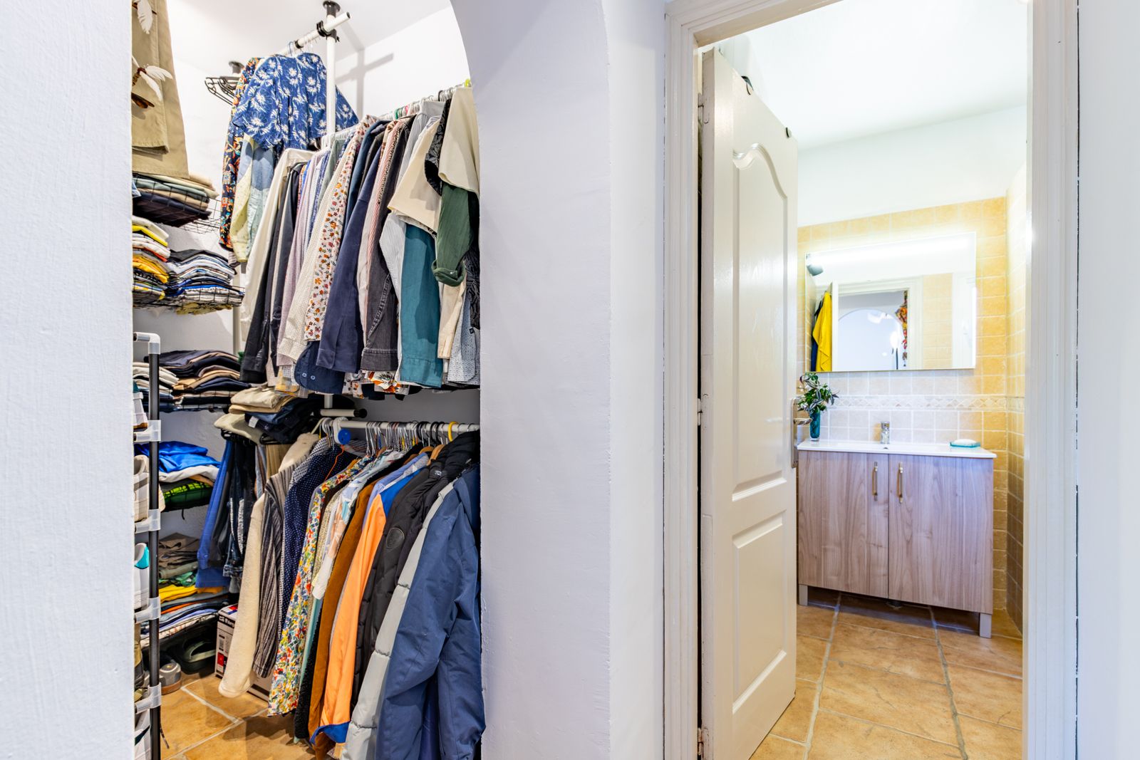 Offener begehbarer Kleiderschrank mit Regalsystem, angrenzend an ein Badezimmer mit Holzwaschtisch, beleuchtetem Spiegel und rustikalen Fliesen.