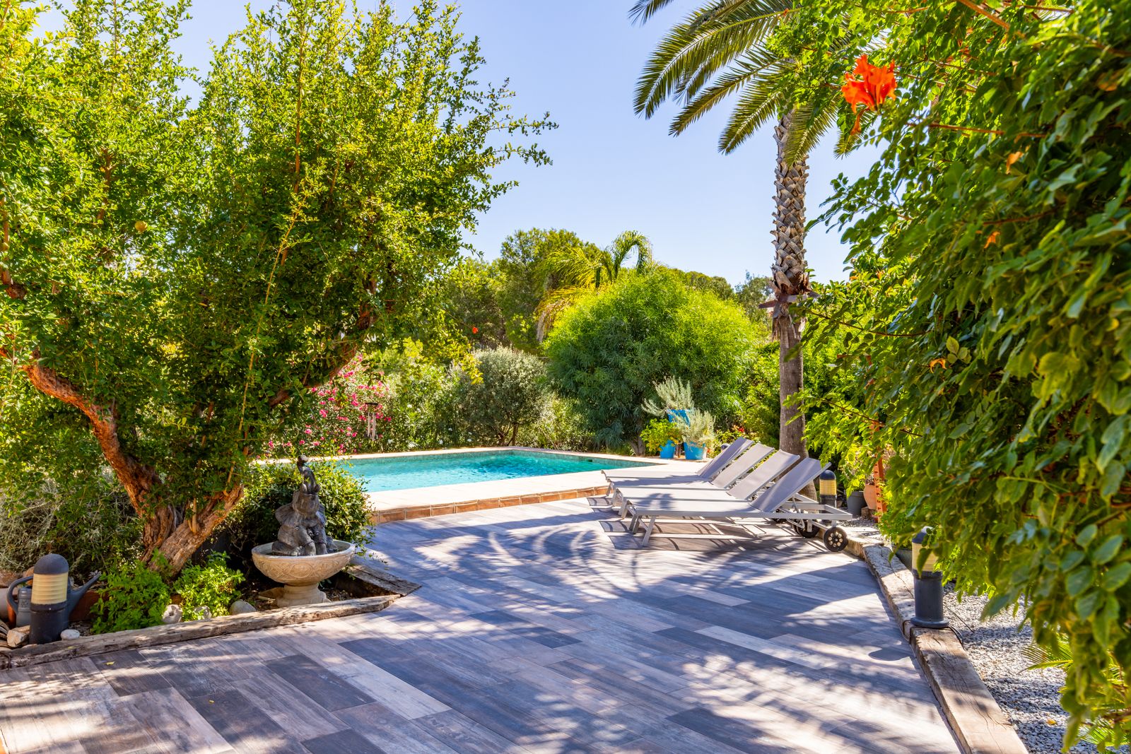 Sonnenterrasse in Holzoptik mit grauen Liegestühlen, umgeben von üppiger mediterraner Bepflanzung, Palmen und einem privaten Swimmingpool.